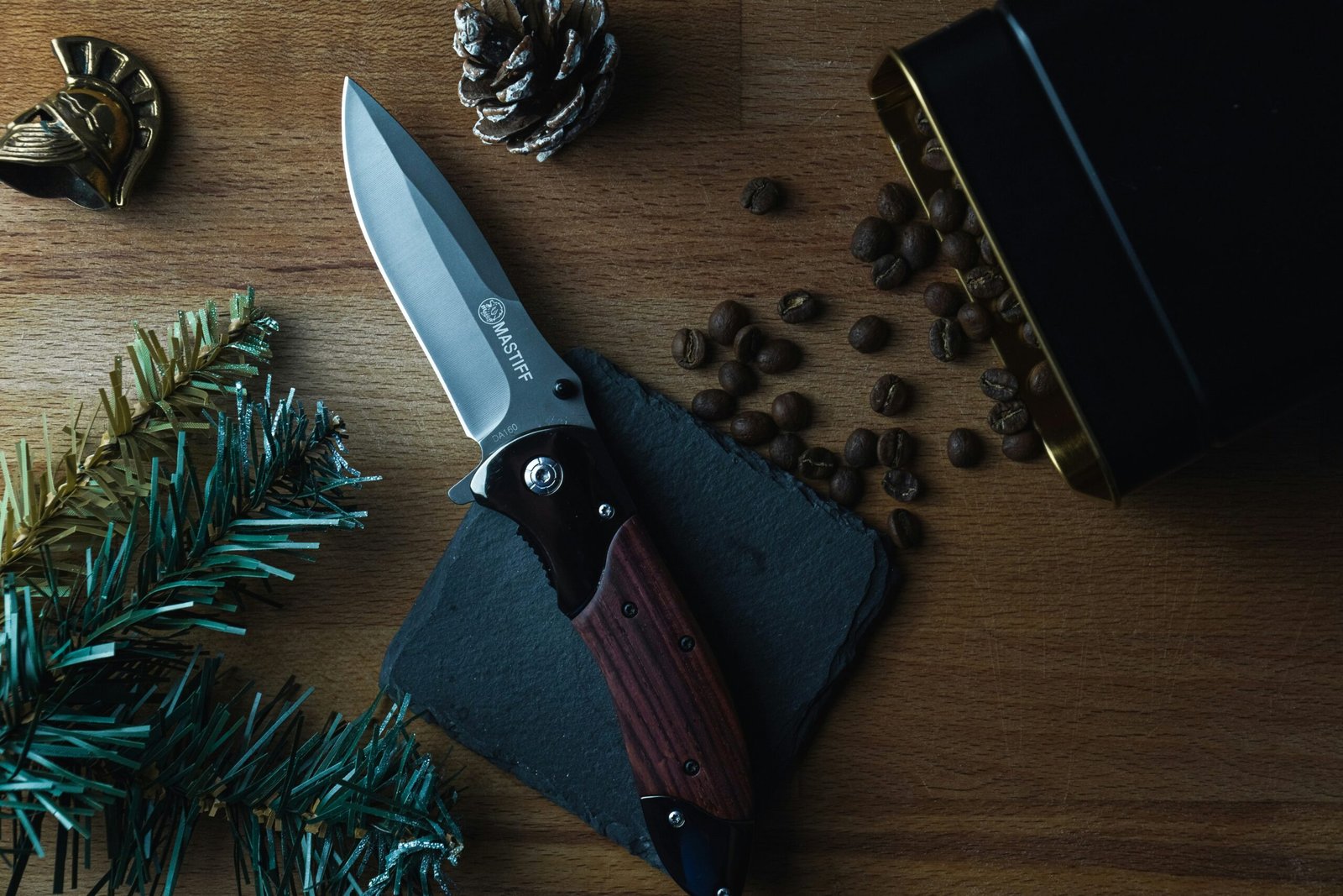 A high-angle, moody shot of a small folding knife with a light wood handle resting on a rustic wooden surface. The handle has the letters engraved on it. A pile of silver thumbtacks is scattered next to the knife.